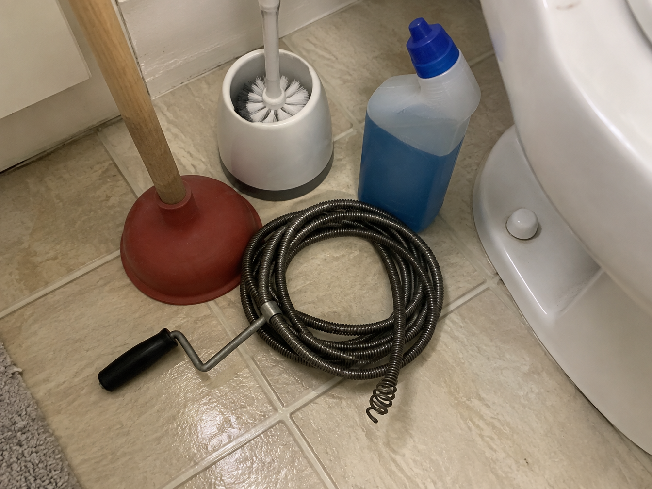 Desatascador de ventosa, sonda, escobilla y gel limpiador amontonados en el suelo del baño junto a un inodoro